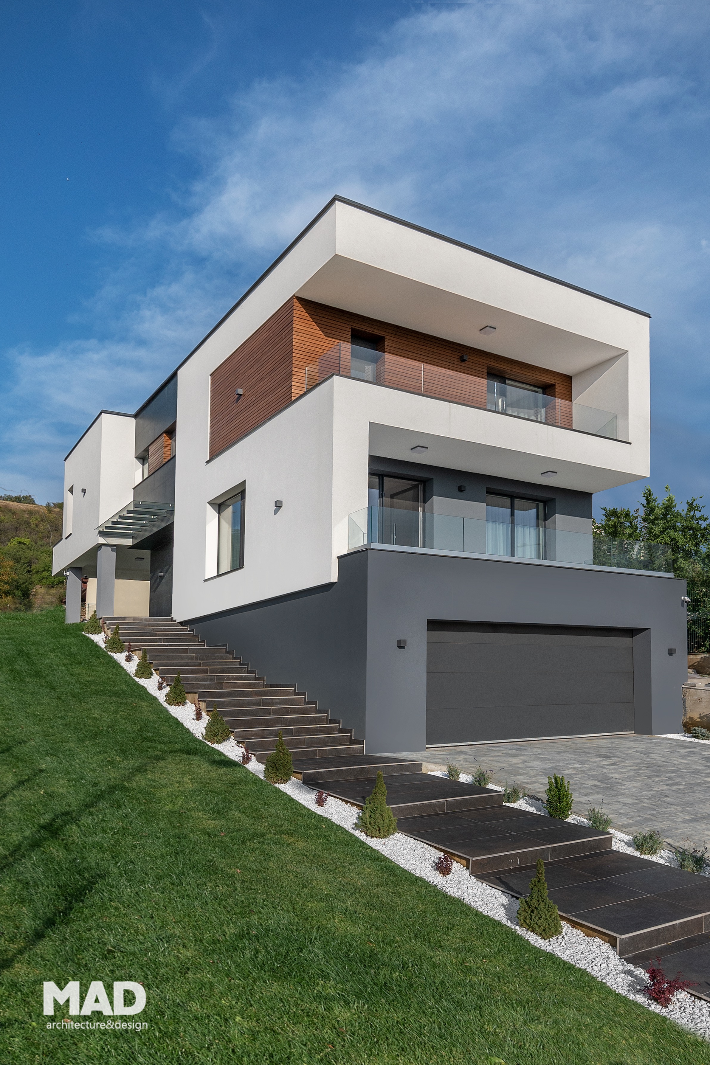 Casă modernă cu demisol, parter și etaj, realizată de MAD architecture&design, fațadă cu tencuială albă și gri antracit, placaj de lemn natural și balustrade din sticlă.