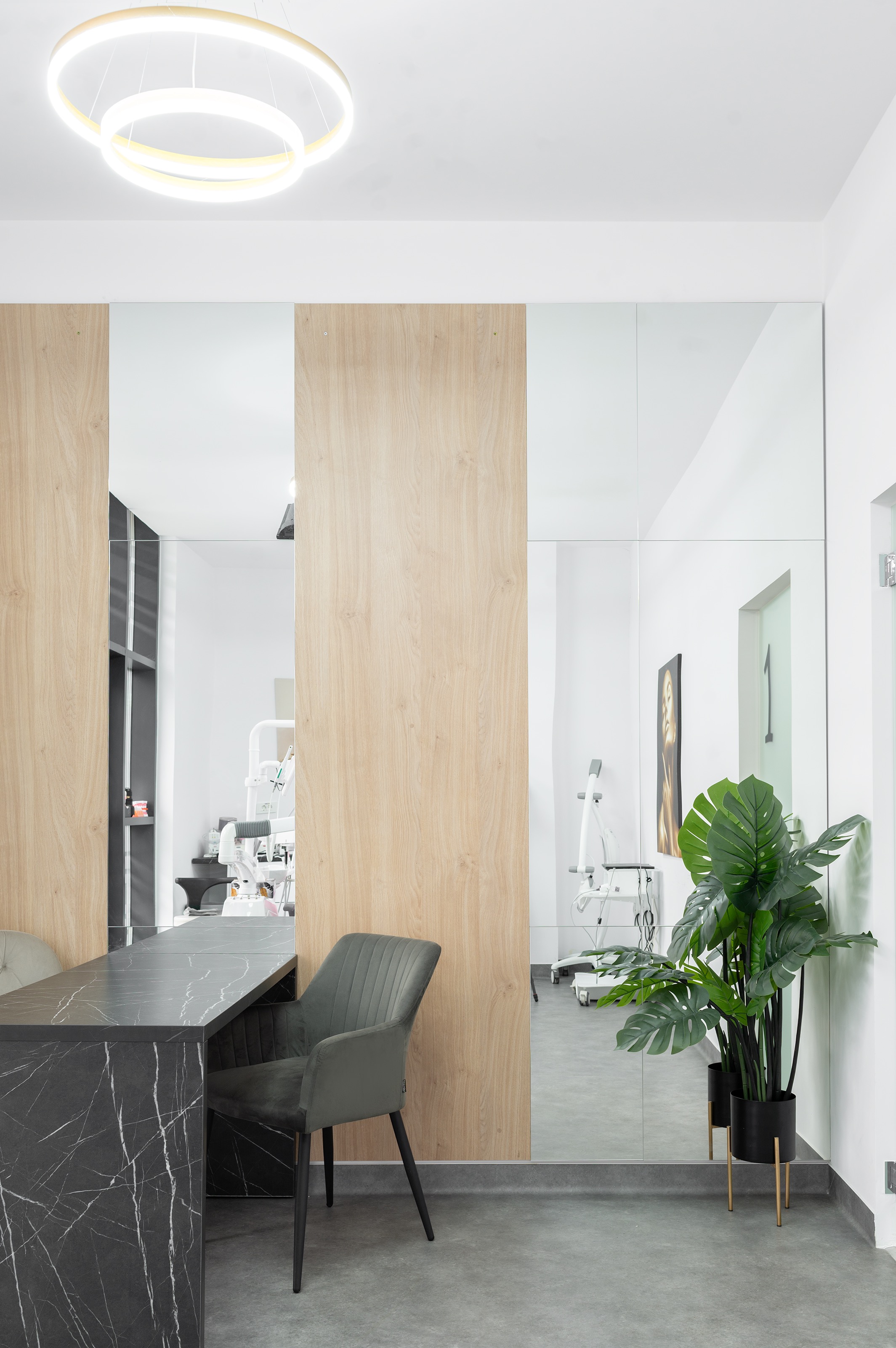 Birou elegant amenajat de MAD architecture&design pentru The Nest Dental Clinic, cu blat PAL cu aspect de marmură neagră, placari cu lemn, oglinzi decorative și scaun tapițat cu catifea gri, planta decorativa exotica monstera in ghiveci, candelabru cercuri aurii LED.