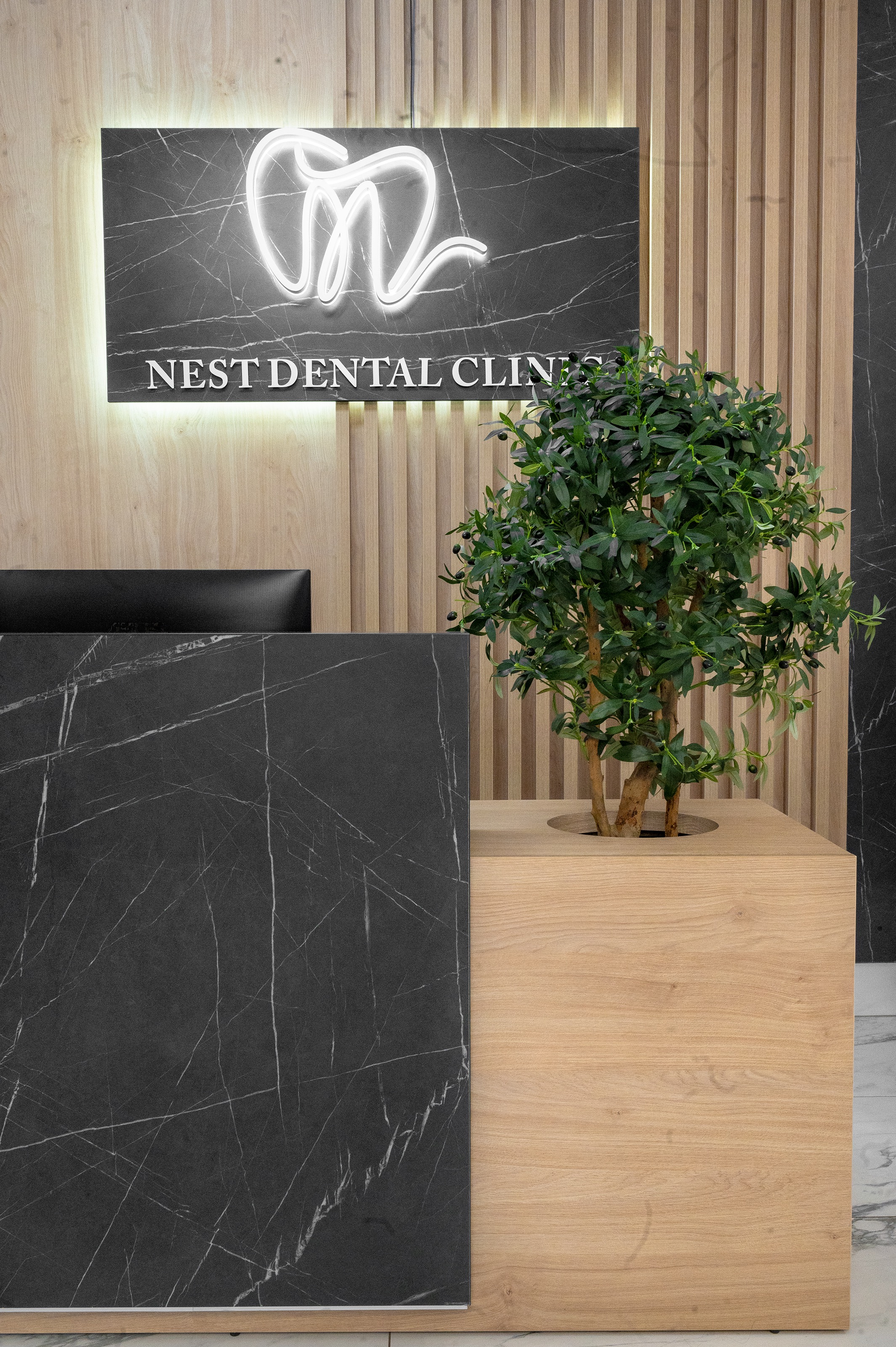 Detaliu al recepției The Nest Dental Clinic proiectată de MAD architecture&design, cu finisaje din lemn natur, panouri negre cu textură de marmură și elemente vegetale integrate in mobilier, un copacel maslin, logo-ul clinicii iluminat cu banda LED.