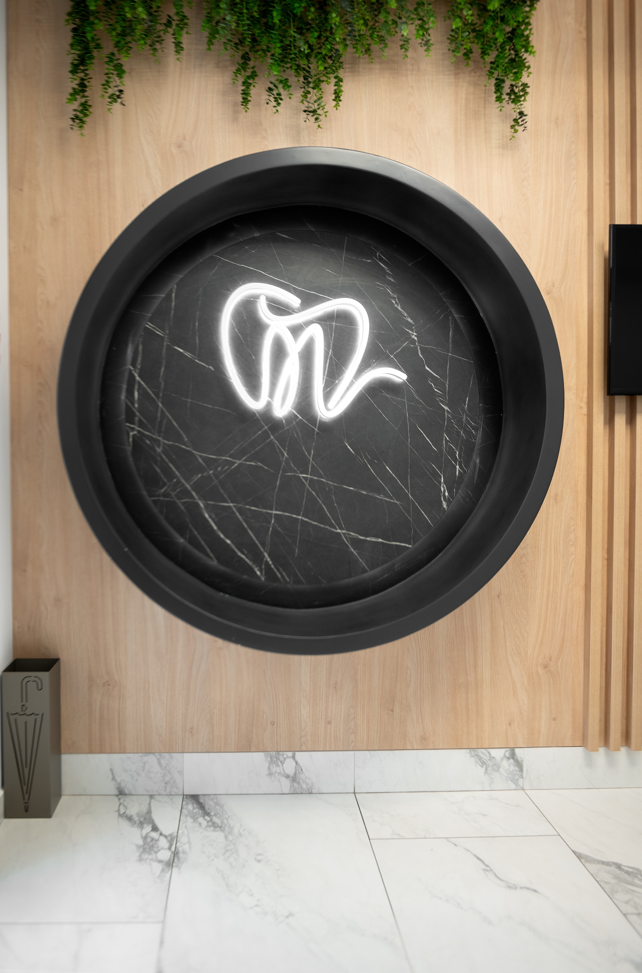 Element decorativ sub forma de cerc cu logo led luminos The Nest Dental Clinic, design realizat de MAD architecture&design, cu fundal din marmura neagră și perete placat cu lemn natur, decorat cu plante curgatoare.