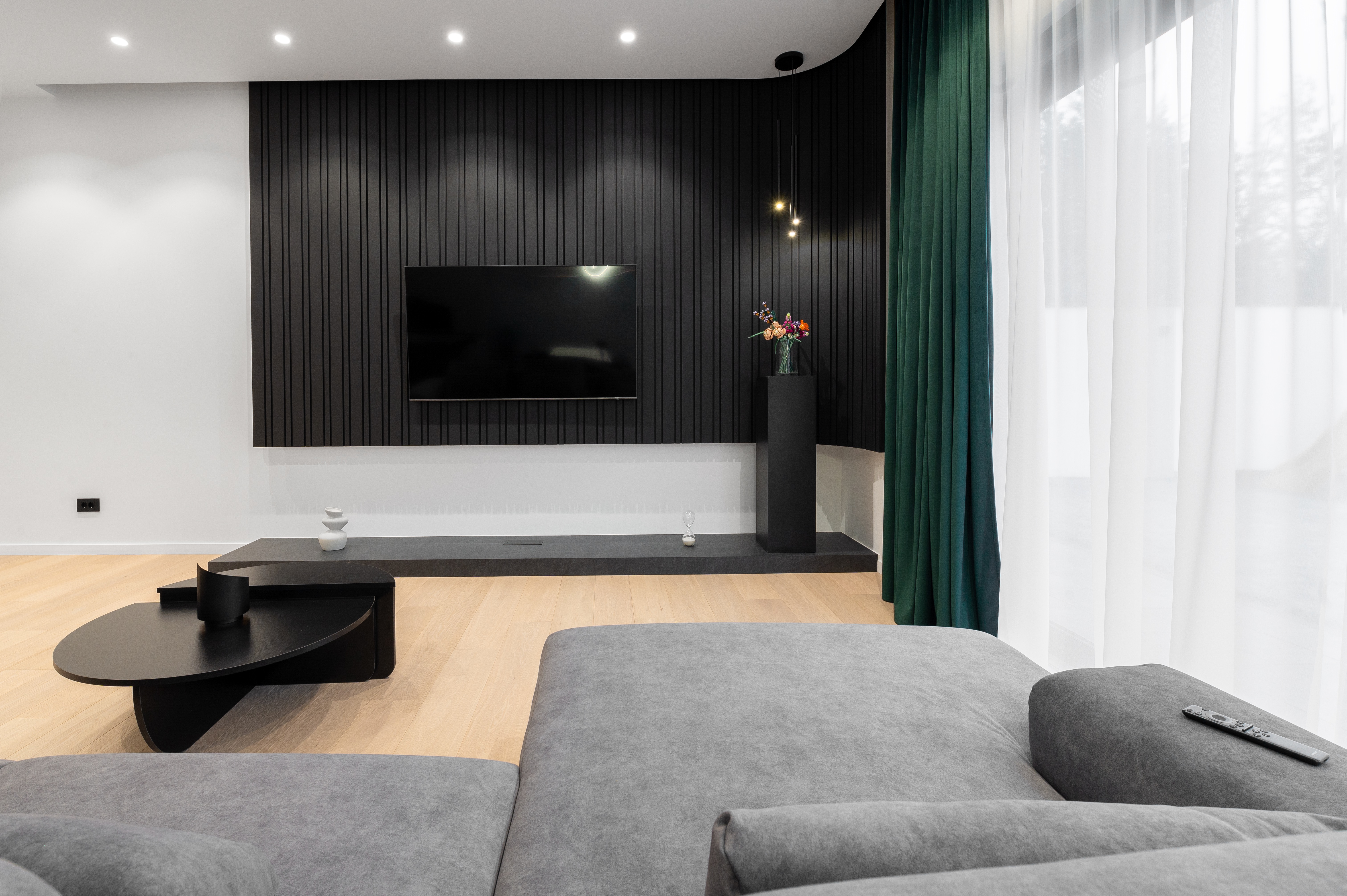 Living contemporan cu canapea gri, panou TV din lamele negre verticale, perdele verzi smarald și accente de lumină caldă – design MAD architecture&design.