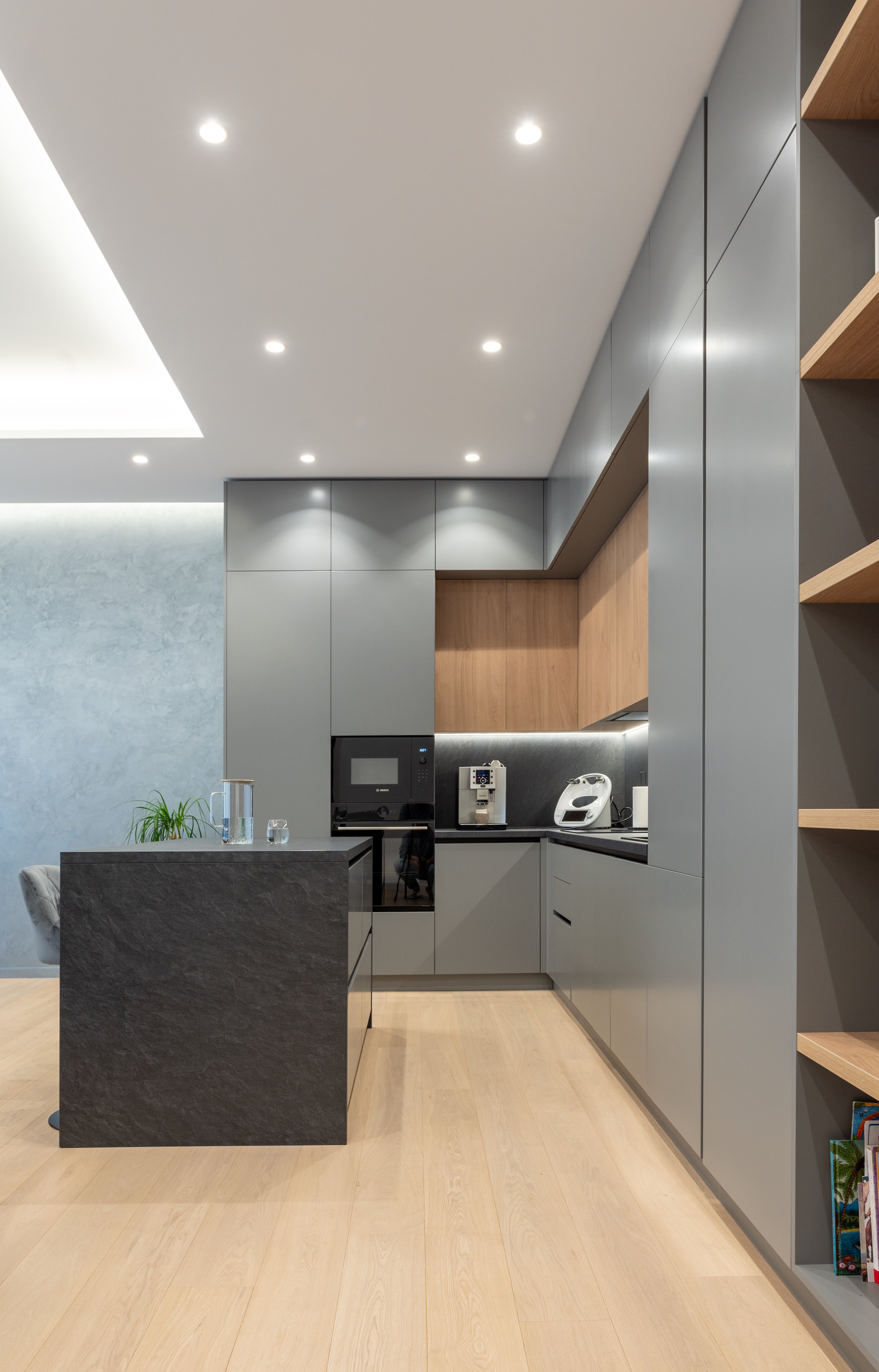 Bucătărie modernă în stil contemporan, cu mobilier gri mat, accente din lemn natural și insulă centrală din piatră neagră; iluminare punctuală în tavan – design semnat MAD architecture&design.