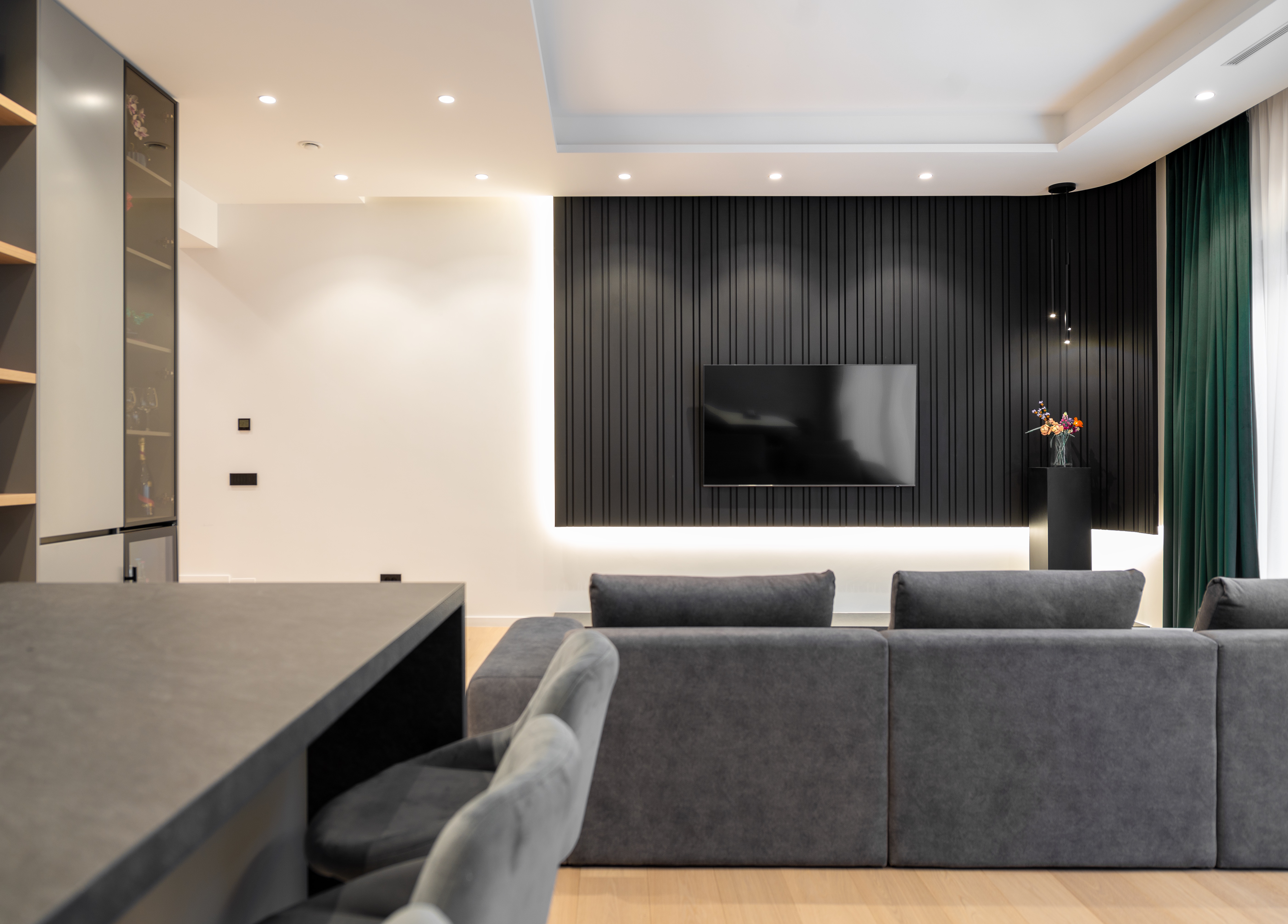 Spațiu open-space contemporan ce integrează livingul și bucătăria, cu canapea gri, insulă de bucătărie din piatră naturală și panou TV negru lamelar – design MAD architecture&design.