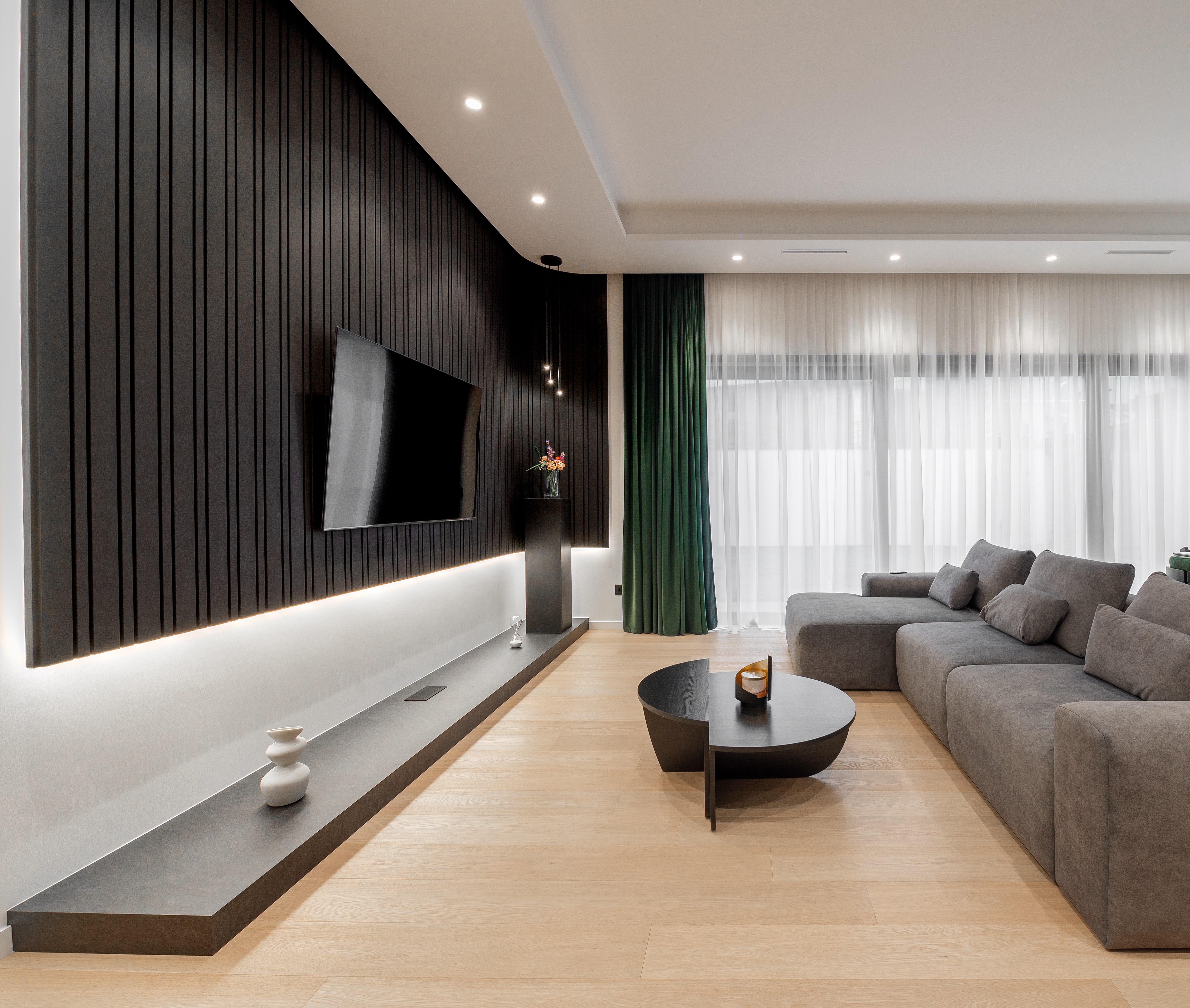 Living modern în tonuri neutre, cu perete TV negru mat, canapea modulară gri și lumini ambientale calde; accente verzi în draperii – proiect MAD architecture&design.