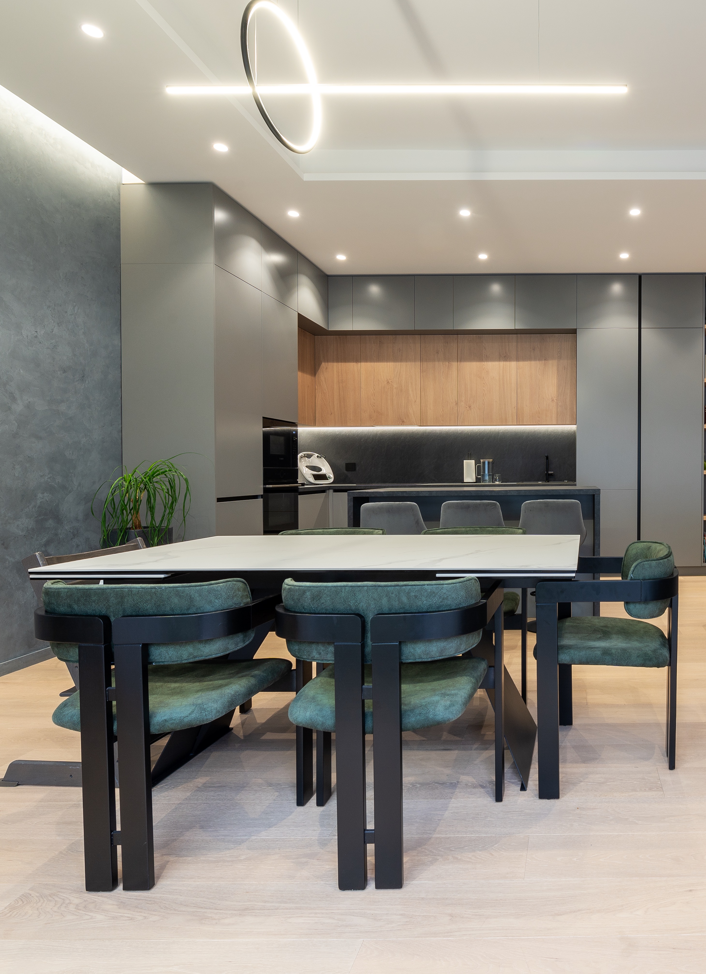 Zonă de dining modernă, cu masă rectangulară albă, scaune tapițate verde smarald și accente metalice negre; bucătăria contemporană se află în fundal – MAD architecture&design.