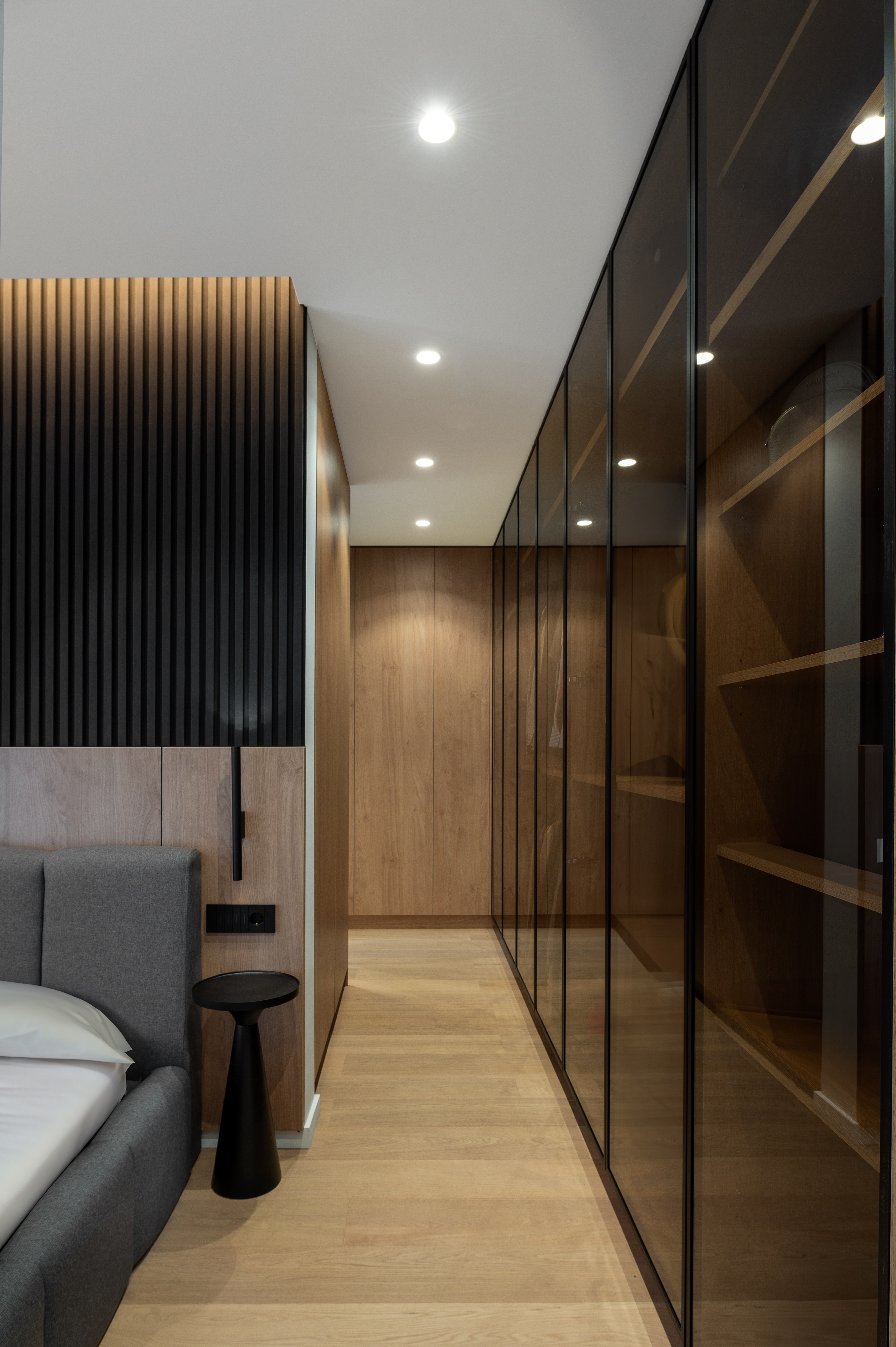 Dormitor matrimonial elegant, cu pat tapițat gri, perete decorativ cu lamele negre și dressing din sticlă fumurie cu rame negre – design MAD architecture&design.