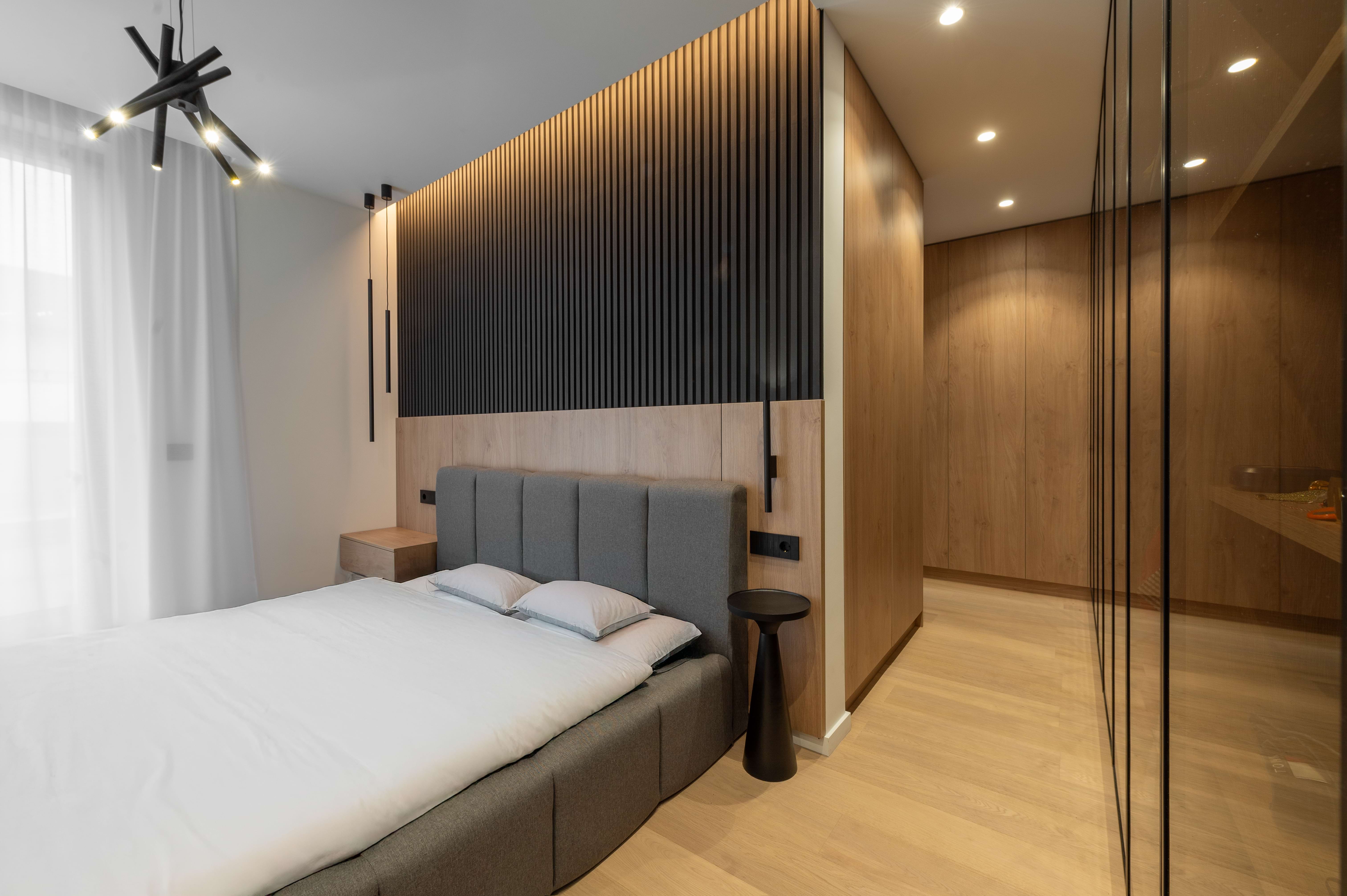 Dormitor matrimonial în stil contemporan, cu pat tapițat gri, perete decorativ din lamele negre verticale și finisaje din lemn natural cald – design MAD architecture&design.