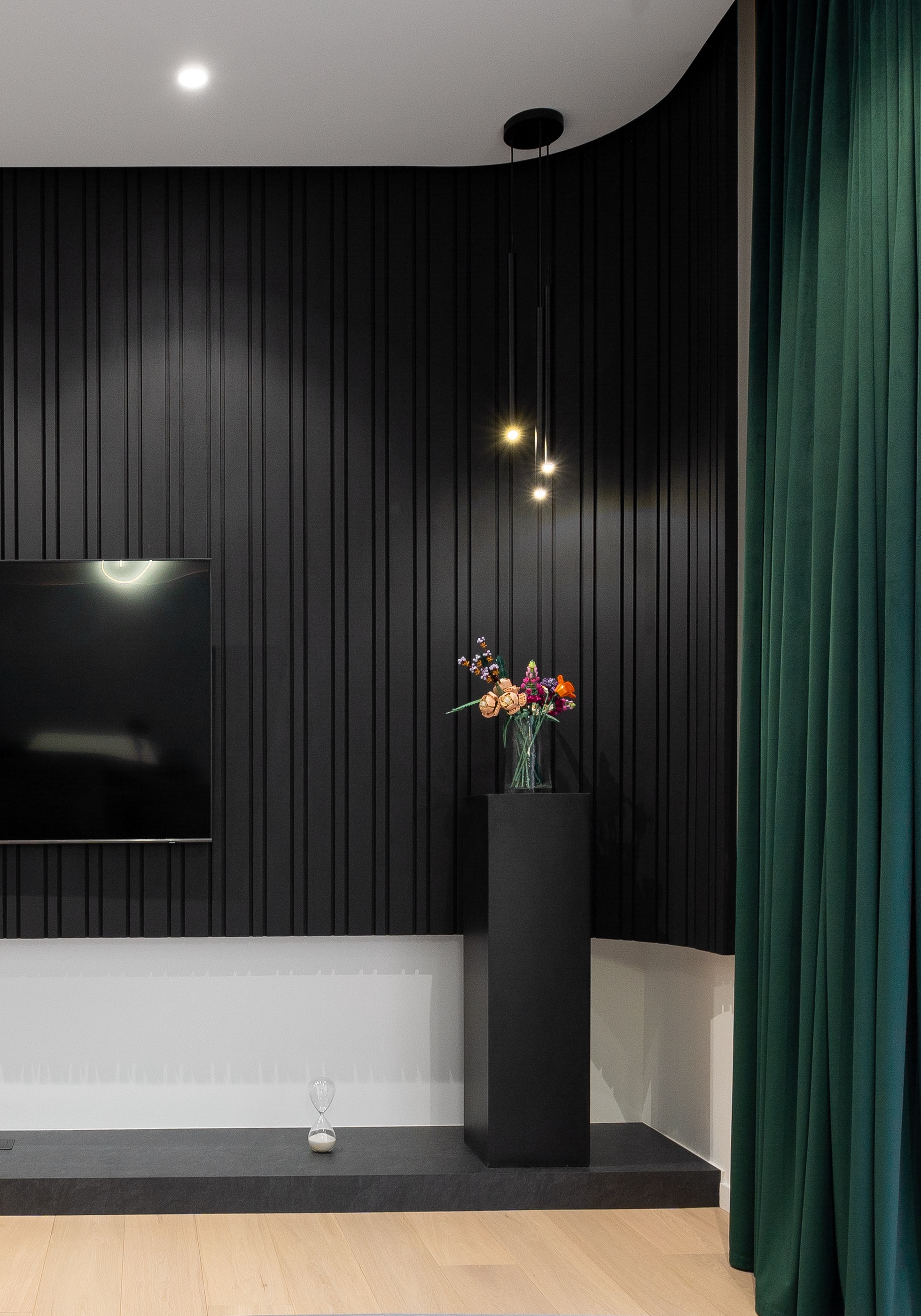 Detaliu din zona de living modern, cu podium decorativ negru mat, panou TV lamelar și piedestal minimalist cu aranjament floral – MAD architecture&design.