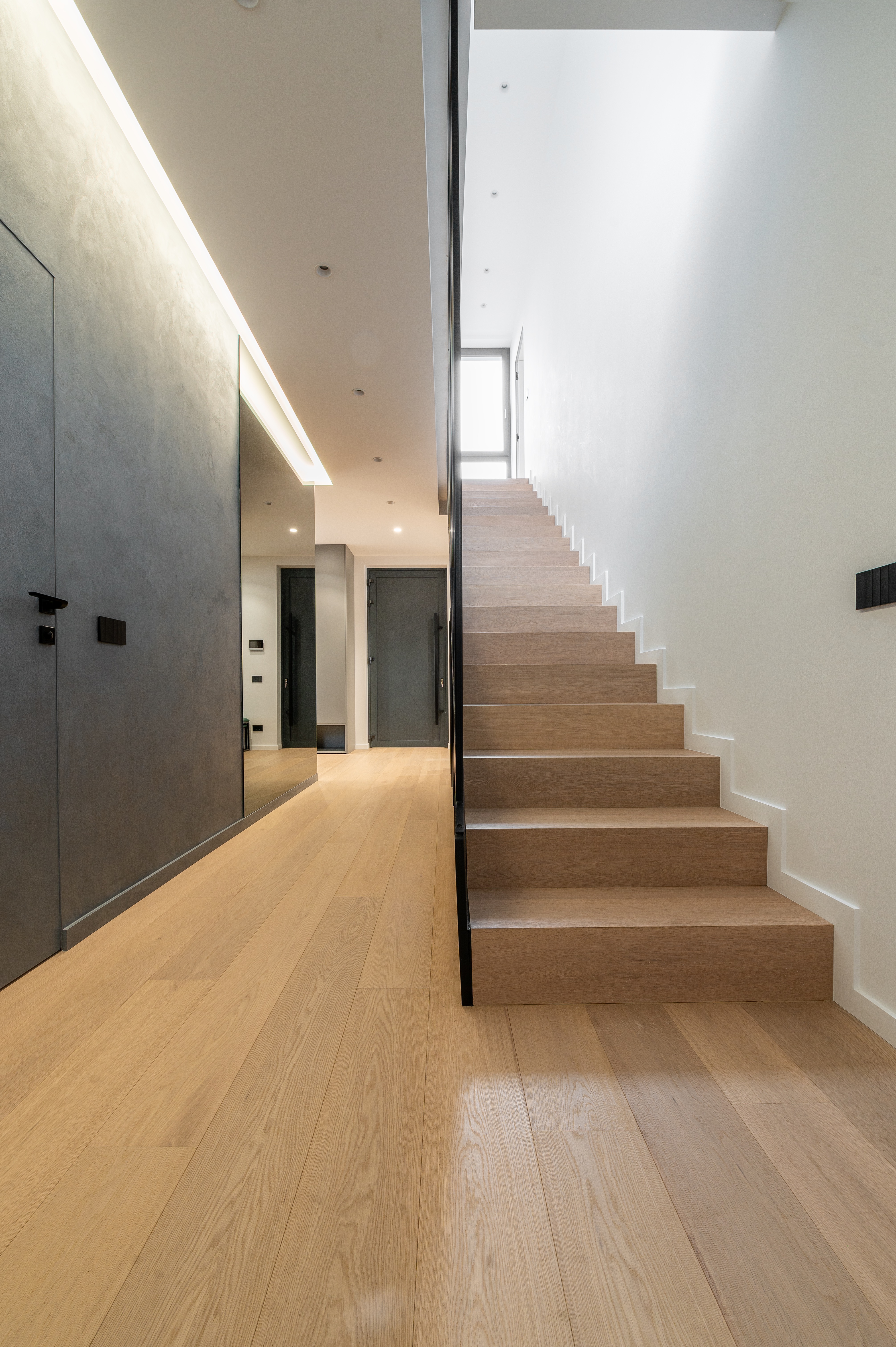 Casă a scării elegantă, cu trepte din lemn deschis, pereți gri texturat și iluminare indirectă în tavan – design contemporan MAD architecture&design.