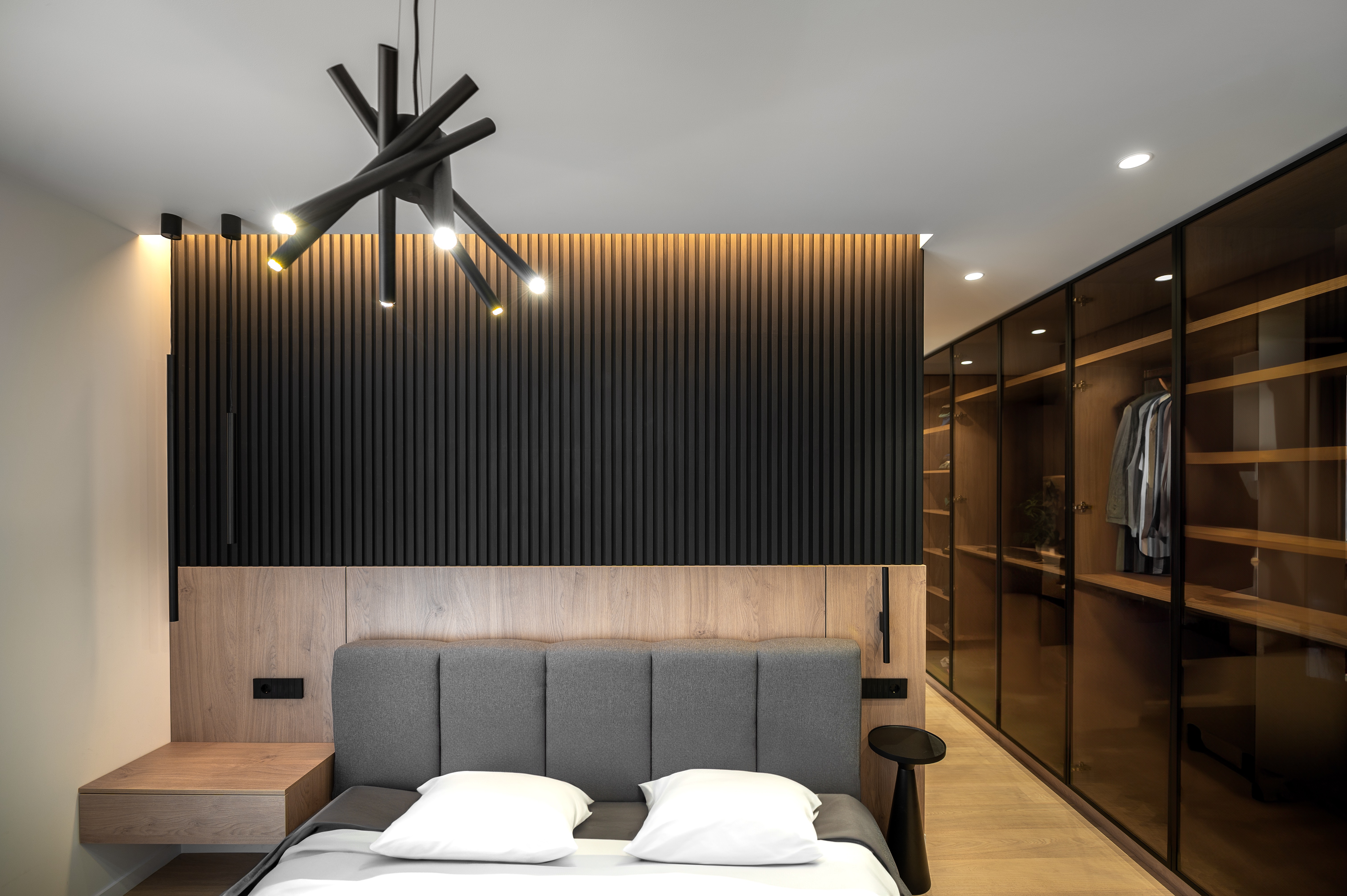 Vedere detaliată asupra dormitorului matrimonial cu tablie tapițată gri, perete din lemn și lamele verticale negre, completat de dressing din sticlă fumurie – MAD architecture&design.